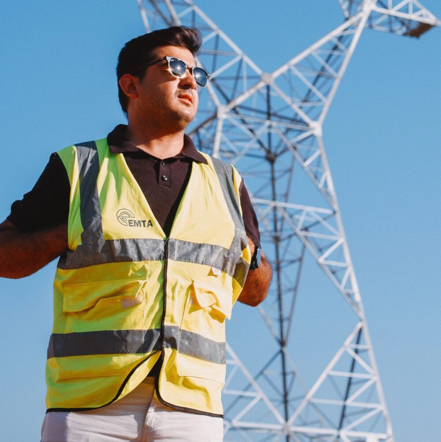 Engineer wearing safety vest inspects towering power lines on a sunny day.
