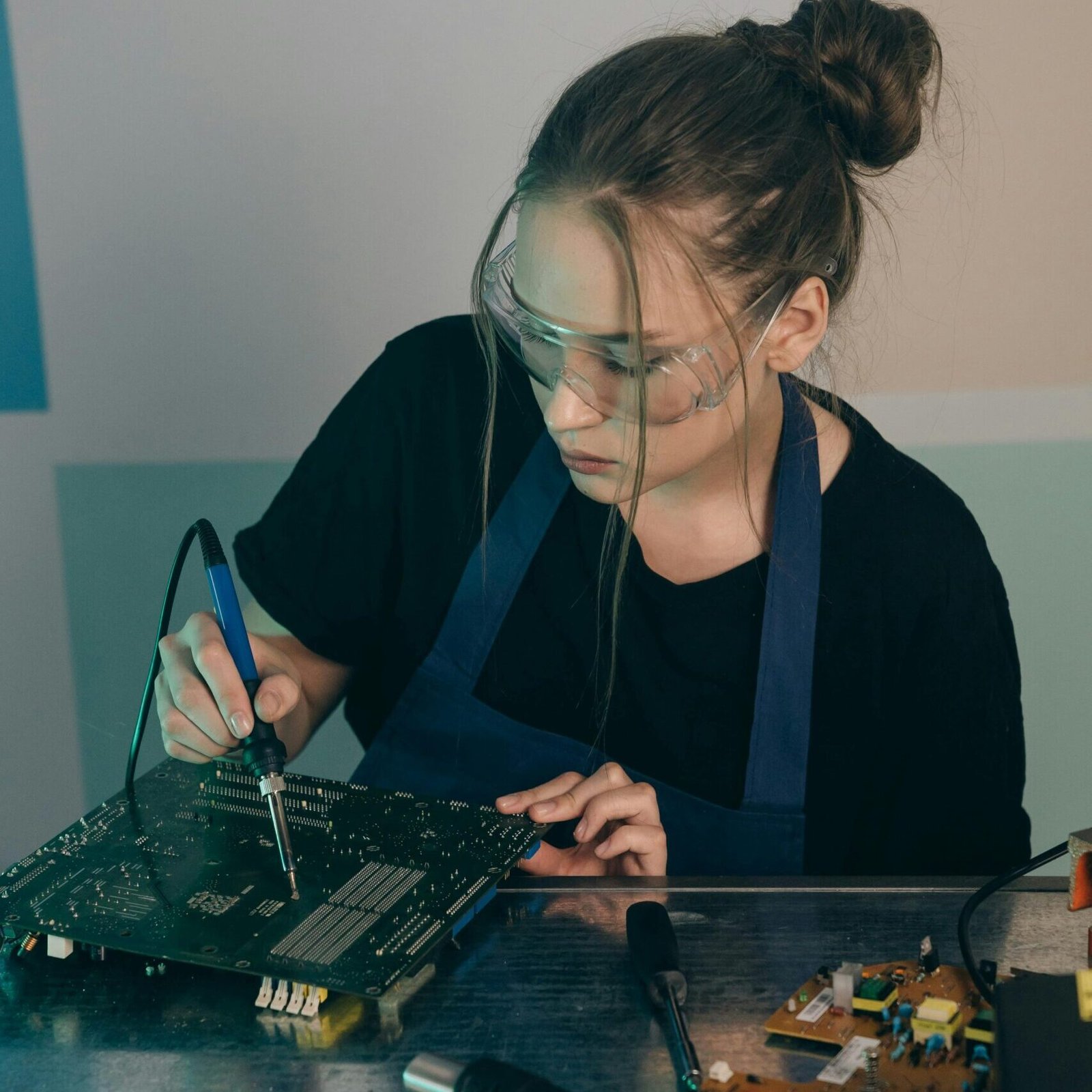 Female engineer wearing goggles soldering an electronic circuit board indoors.